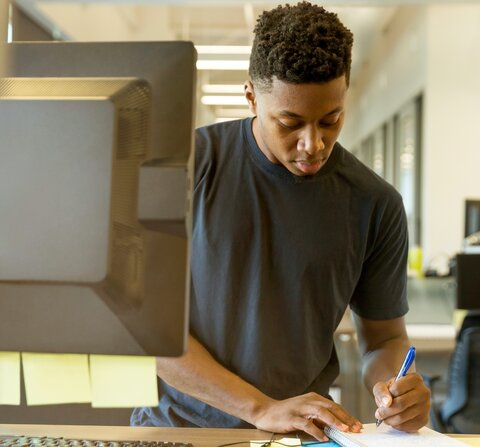 A person is standing in front of a computer. They are writing something on a small notepad.