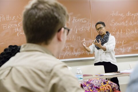 A professor of English teaches next to a brown chalk board.
