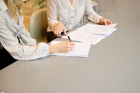 Two people from the chest down are pictured. They are looking at three piles of career-related papers like resumes and CVs.