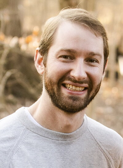Brandon is wearing a light grey sweater. He is smiling directly at the camera.