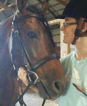 Katie is pictured with one of the horses under her care. Katie wears glasses, a horse-riding helmet, and a turquoise shirt. The horse is brown.