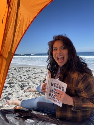 Hilary smiles at the beach. She is holding a book titled Heads of the Colored People.