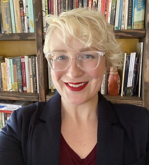 Lesley smiles in front of bookshelves with many books. Lesley is wearing clear glasses and a navy blazer.