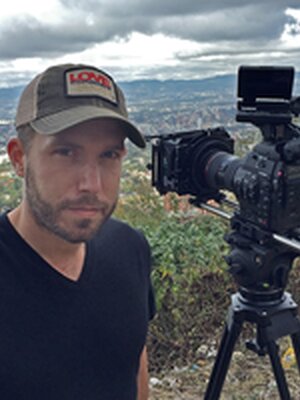 Ryan smiles standing next to a camera on a tripod. He is wearing a hat and a black v-neck shirt.