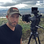 Ryan smiles standing next to a camera on a tripod. He is wearing a hat and a black v-neck shirt.